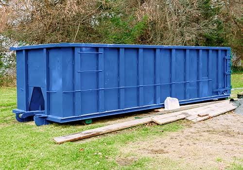 A blue roll-off dumpster in a yard next to wood scraps and lumber